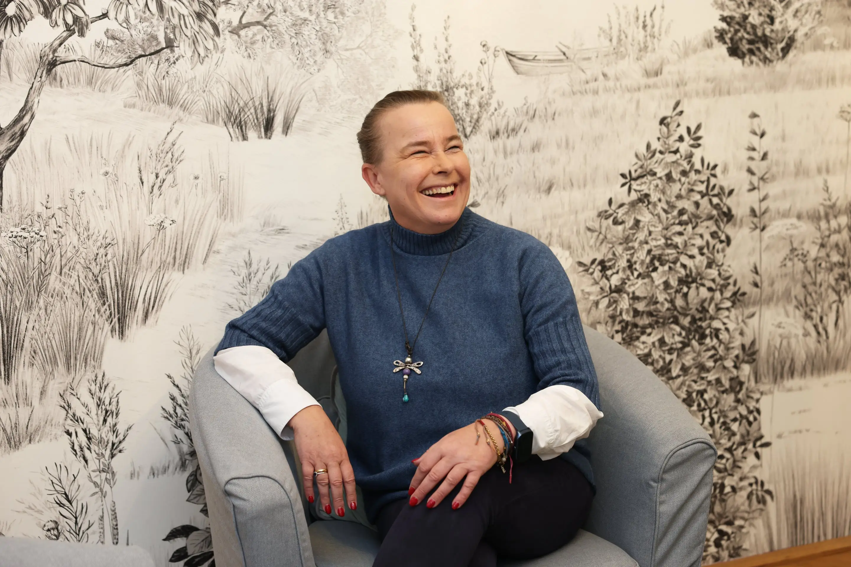 Verena Broschek-Fellner lacht in einem Sessel vor der gezeichneten Landschaftstapete im Glücksraum